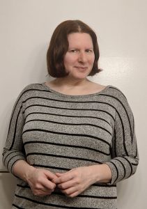 Rachel Donner 1 Image of Rachel Donner. She is wearing a black and grey striped shirt and is standing against a white background. She has shoulder length brown hair and is looking slightly sideways, smiling.